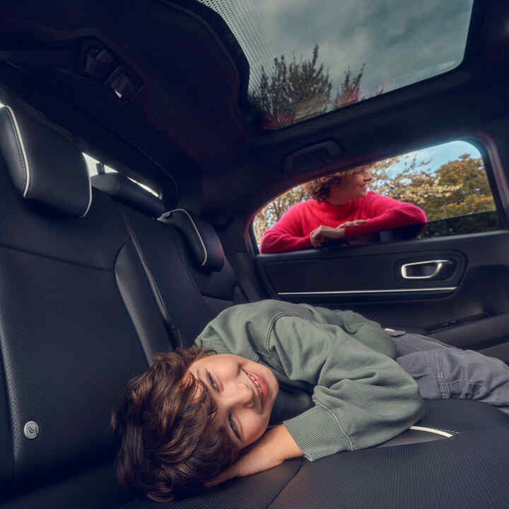 Kind auf dem Rücksitz eines Honda e:Ny1 liegend, die Mutter steht vor dem Fenster.