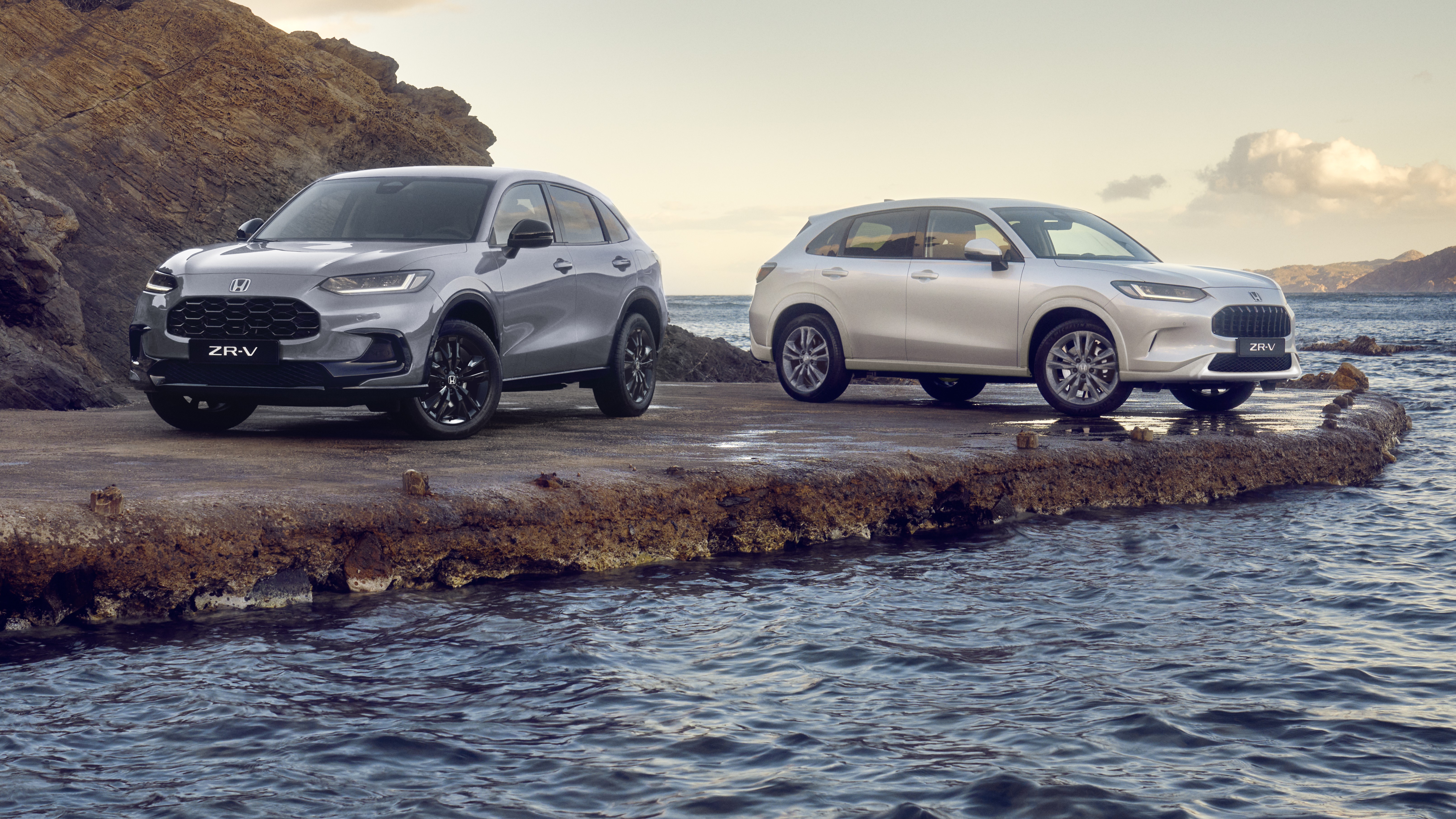 Zwei Honda ZR-V SUVs, einer in Blau und einer in Weiß, stehen auf einem felsigen Küstenvorsprung. Im Hintergrund sind Wasser und Hügel unter einem leicht bewölkten Himmel zu sehen.