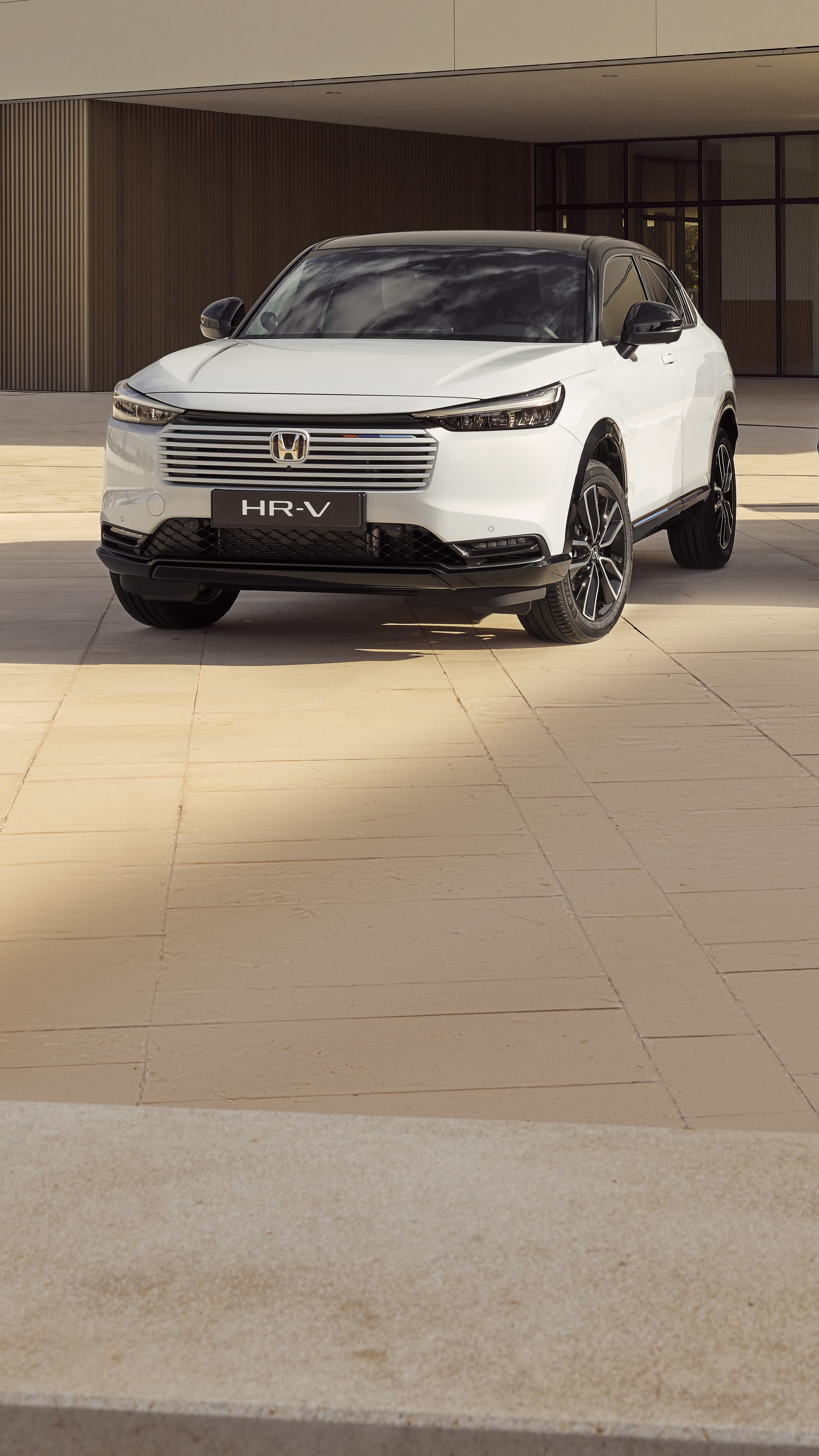 Ein weißer Honda HR-V SUV vor einem modernen Gebäude mit beige gefliesten Wänden geparkt. Das Fahrzeug ist schräg positioniert und zeigt sein elegantes Design sowie den markanten Kühlergrill.