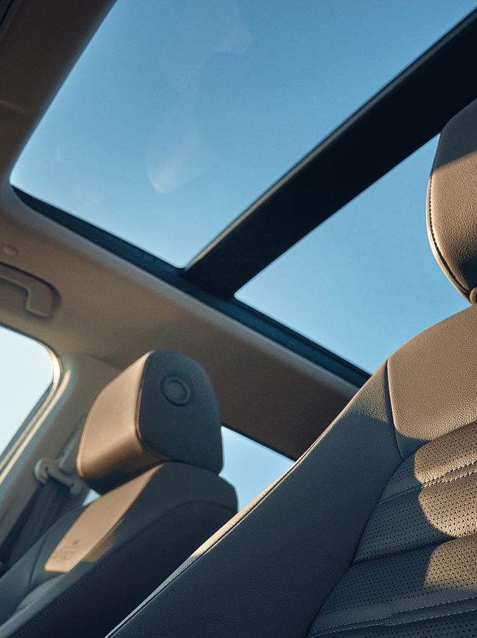 Innenansicht eines Autos mit Panorama-Glasschiebedach, das den blauen Himmel und Sonnenlicht hereinlässt, sowie mit Ledervordersitzen und Kopfstützen im vorderen Bereich, die von natürlichem Licht erhellt werden.