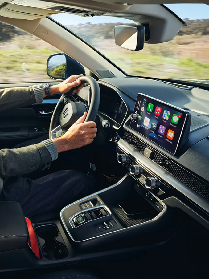 Innenansicht eines modernen Autos mit Fahrer hinter dem Steuer, sichtbar sind Armaturenbrett, Lenkrad, Mittelkonsole und ein großer Touchscreen mit farbigen App-Symbolen. Außerhalb zieht die Landschaft vorbei.