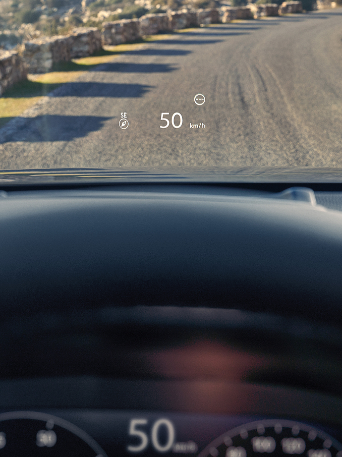 Eine Perspektive vom Fahrersitz zeigt ein Head-Up-Display (HUD), das „50 km/h“ und einen Kompass auf die Windschutzscheibe über einer Schotterstraße projiziert.