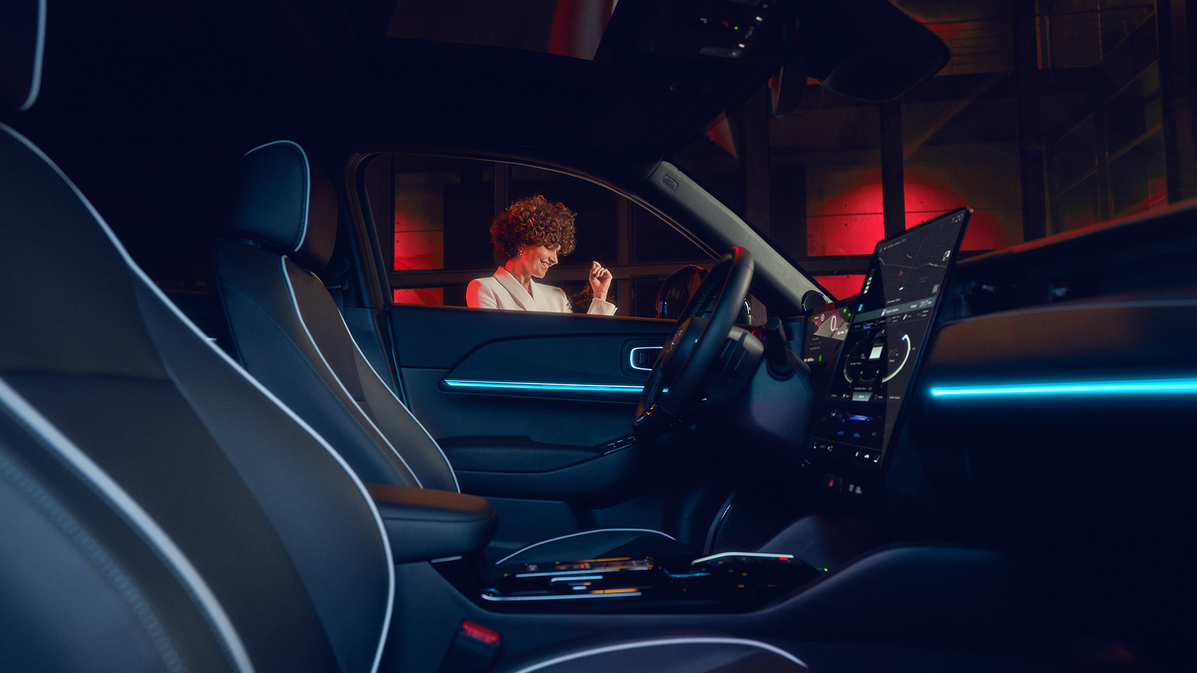 Honda e:Ny1, beleuchteter Innenraum bei Nacht mit Model vor dem Fenster.