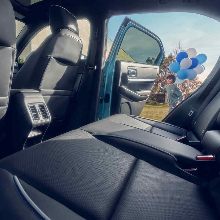 Honda e:Ny1, Nahaufnahme des Rücksitzes und ein Kind, das mit Luftballons an der offenen Tür vorbeigeht.