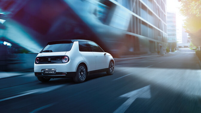 Honda e, 3/4-Frontansicht, beim Fahren durch eine elektrisierte Stadt.