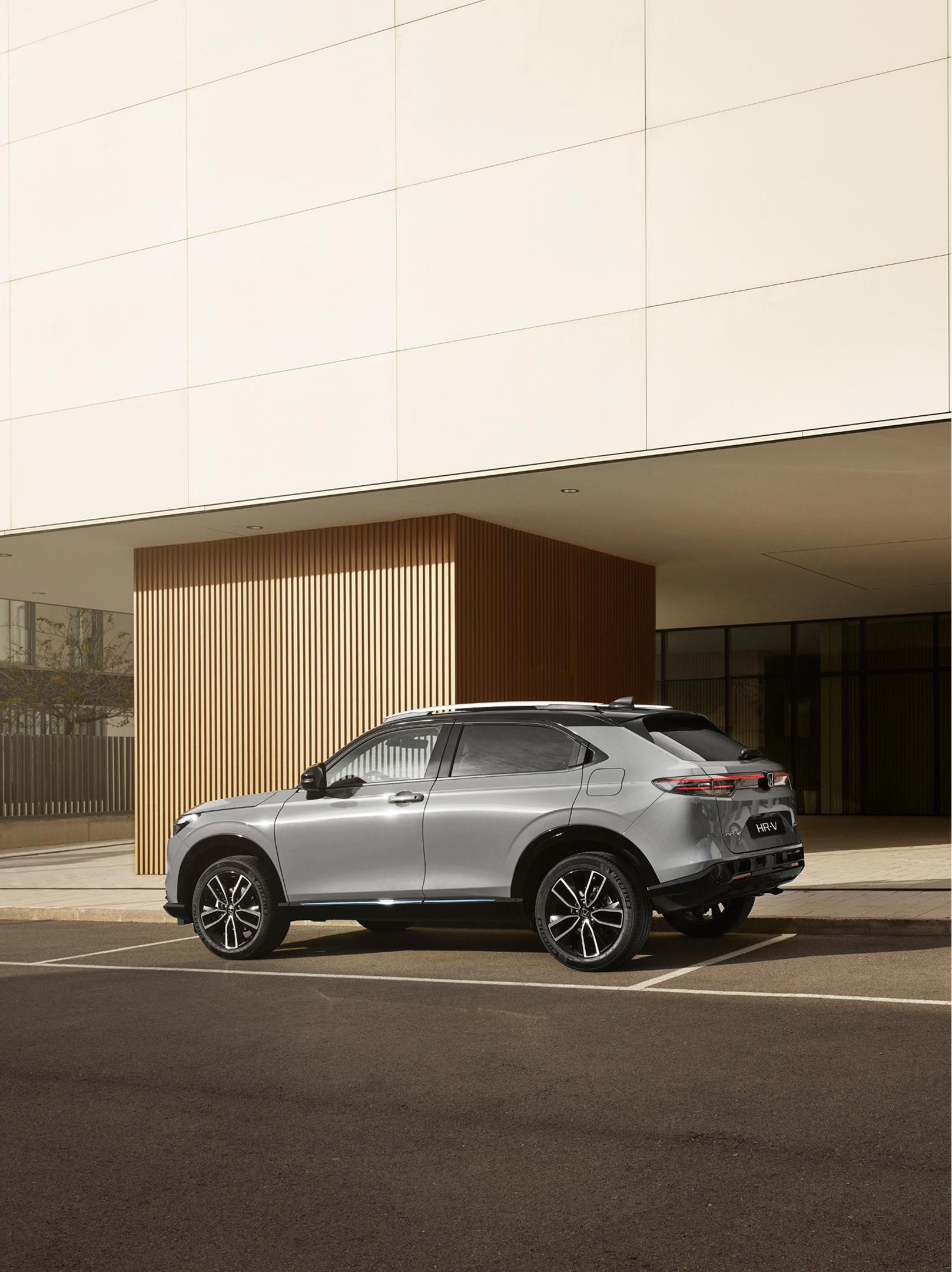 Ein silberner Honda HR-V steht auf einem Außenparkplatz vor einem modernen Gebäude mit Glaswänden und vertikaler Holzverkleidung, aus einem hinteren Dreiviertelwinkel bei Tageslicht fotografiert.
