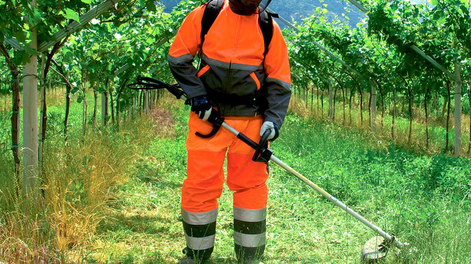 Modell mit rückentragbarem Freischneider auf einem Feld.