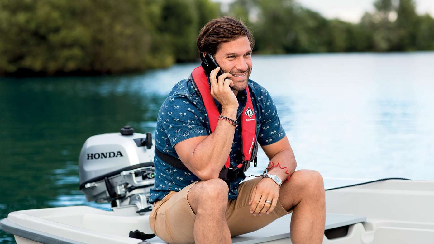 Mann auf kleinem Boot, der während der Unterhaltung sein Telefon am Honda Außenbordmotor auflädt