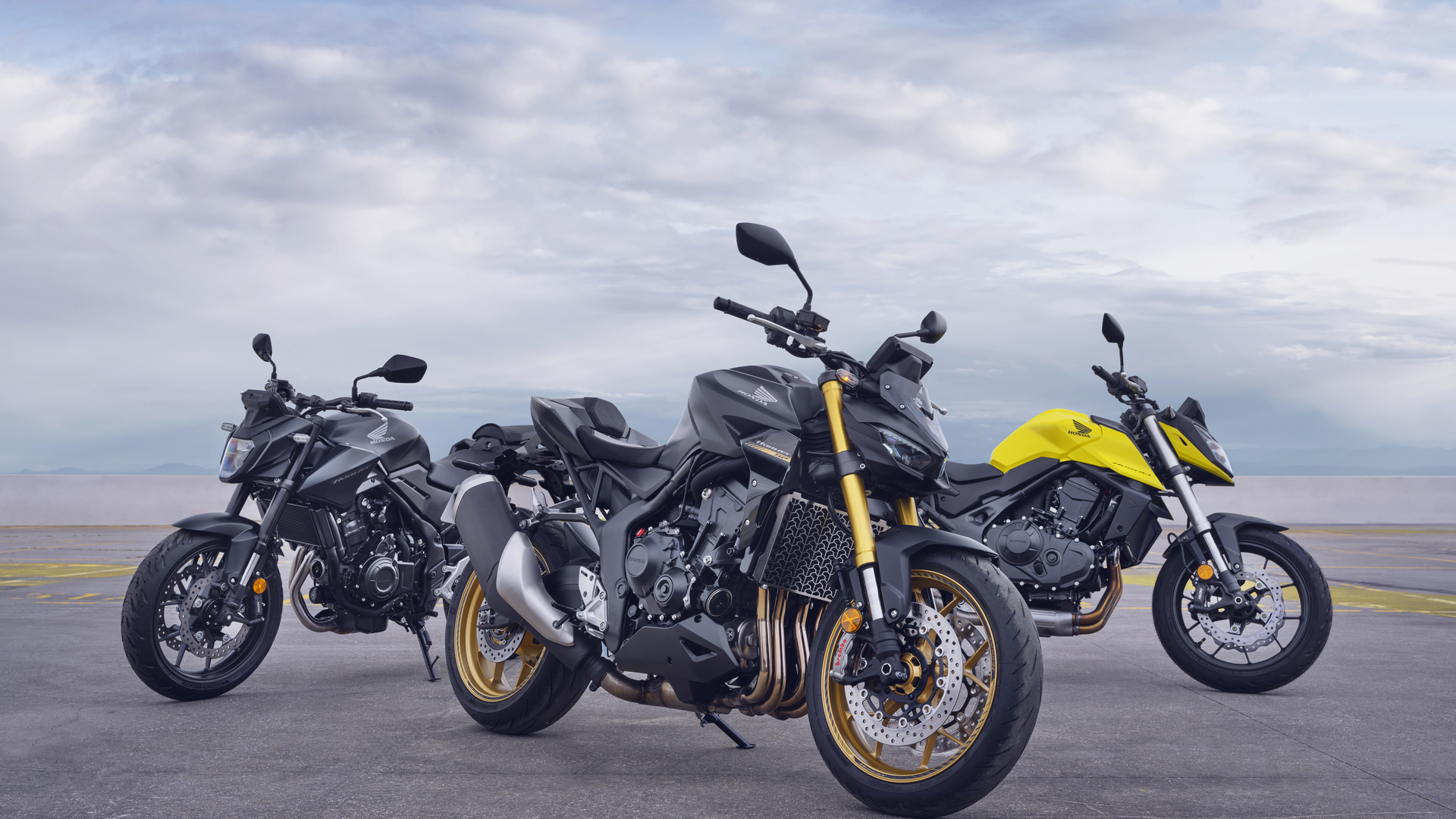 Three Honda CB650R motorcycles lined up on a paved surface against a cloudy sky. The bikes are in different colors: black, gray, and yellow, showcasing the model's sporty naked bike design with distinctive round headlights and muscular fuel tanks.