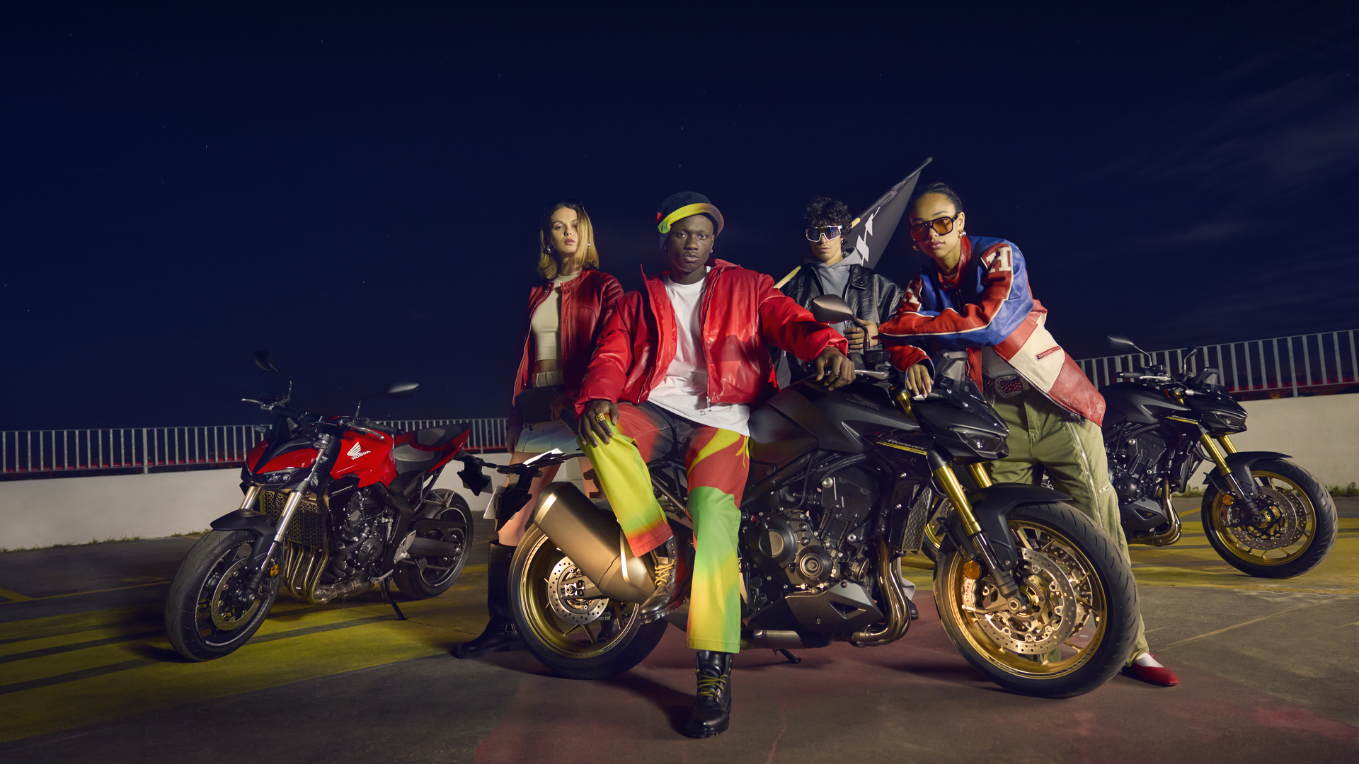 Four people in colorful outfits pose with Honda motorcycles at night, including models like the CB1000R and CB650R. The riders are positioned around their bikes on a dimly lit outdoor surface.