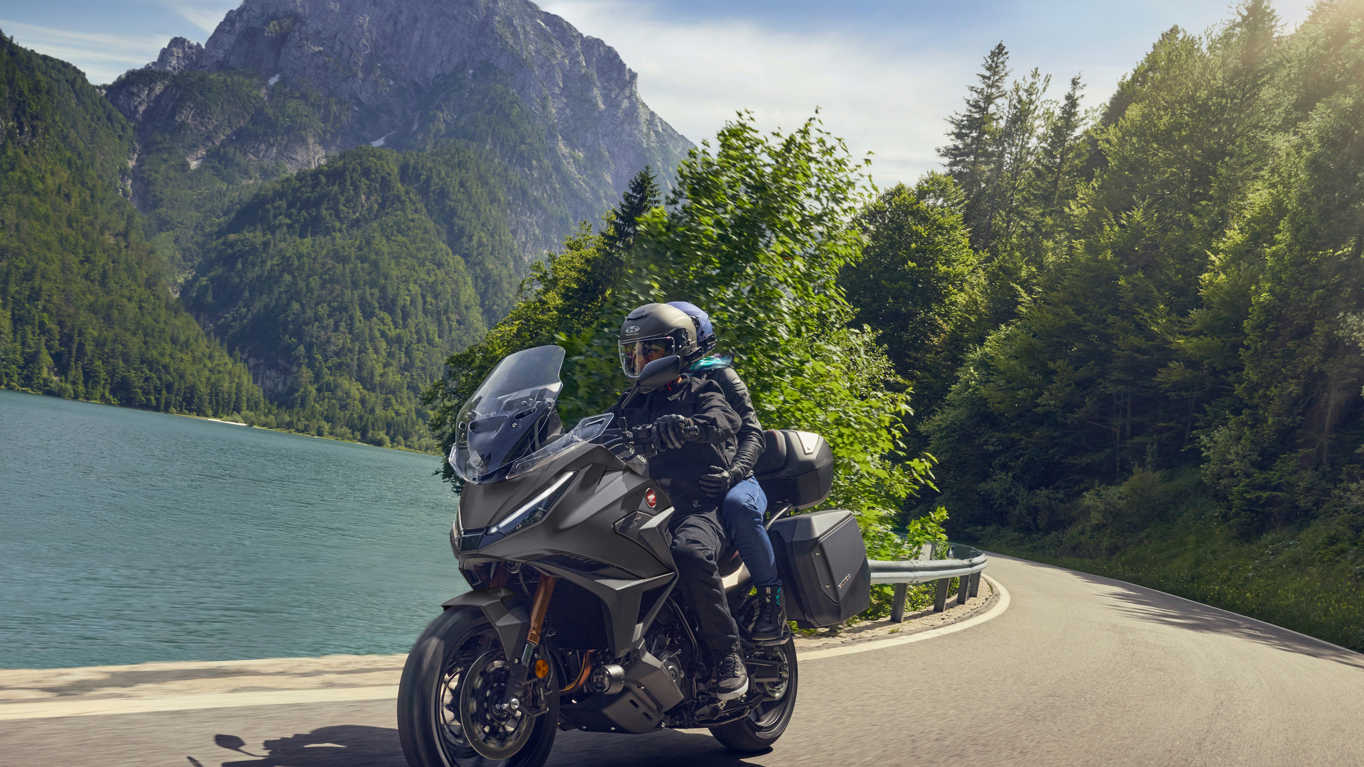 A Honda NT1100 motorcycle being ridden along a winding mountain road beside a turquoise lake. The rider, wearing dark gear and helmet, leans into a curve with forested mountains in the background.