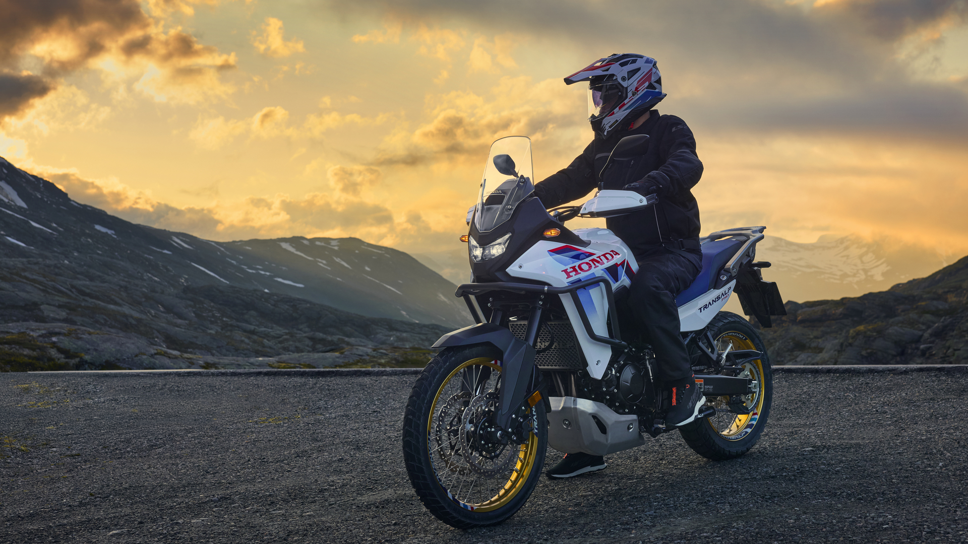 A Honda Africa Twin adventure motorcycle parked on a mountain road at sunset. The rider, wearing a black helmet and jacket, sits astride the bike against a backdrop of dramatic cloudy sky and distant mountains.