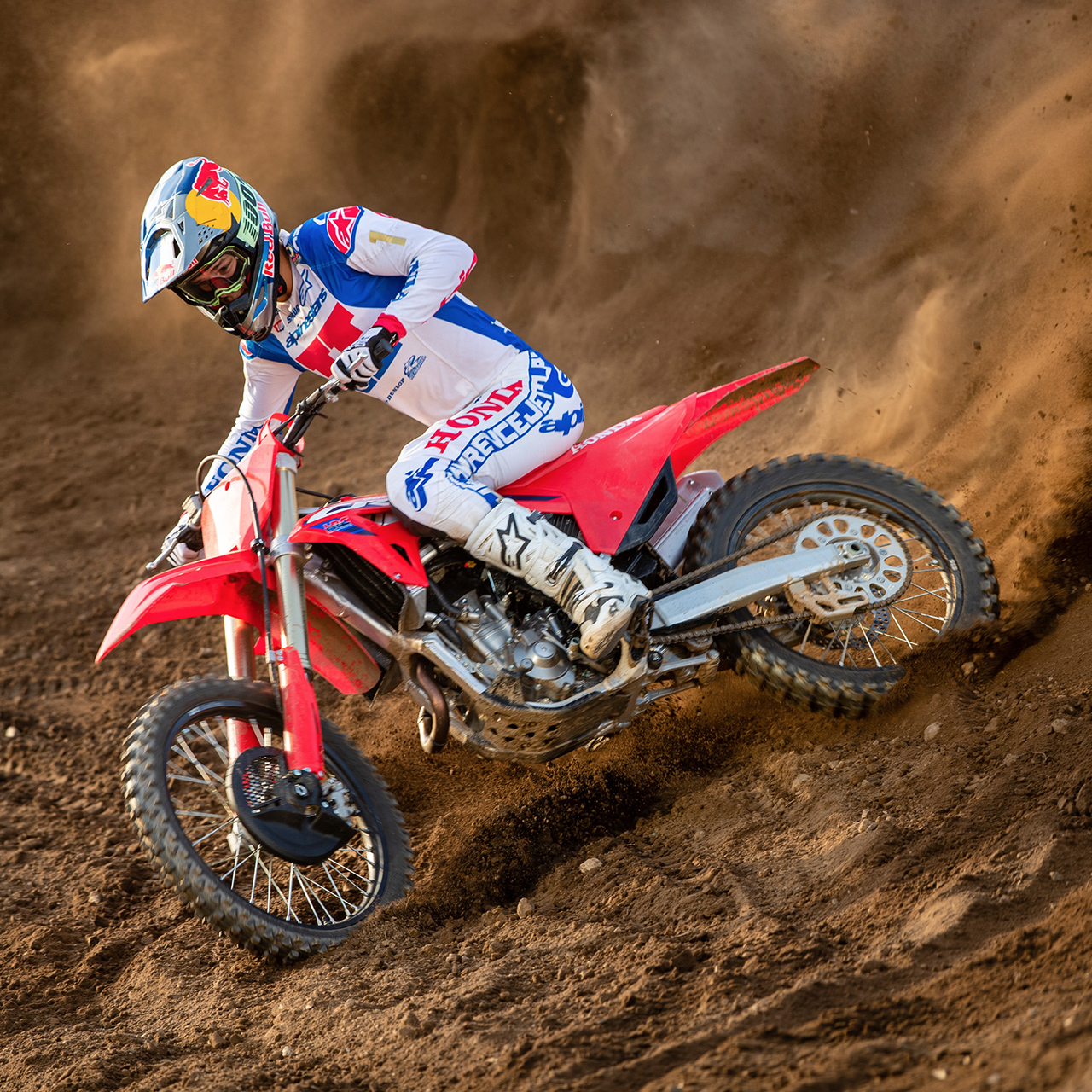 Ein Motocross-Fahrer in weißer, blauer und roter Rennkleidung legt sein rotes Dirt-Motorrad scharf in eine Kurve, durchpflügt eine sandige Strecke und wirbelt dabei eine beeindruckende Staubwolke hinter sich auf.