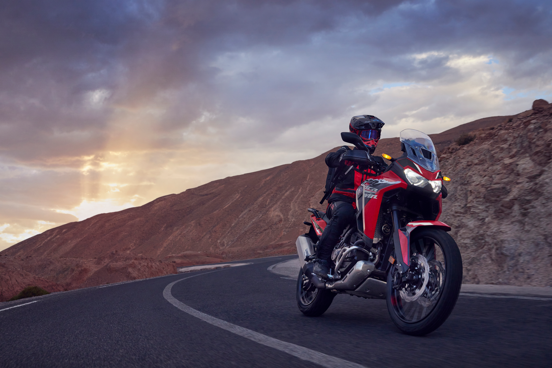 Rote Africa Twin mit Fahrer auf einer asphaltierten Straße mit Bergen und einer untergehenden Sonne im Hintergrund