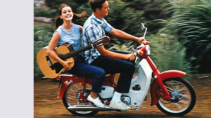 Honda Cub mit Fahrer und Beifahrerin mit Gitarre. Auf einer unbefestigten Straße. Aufnahme von rechts.