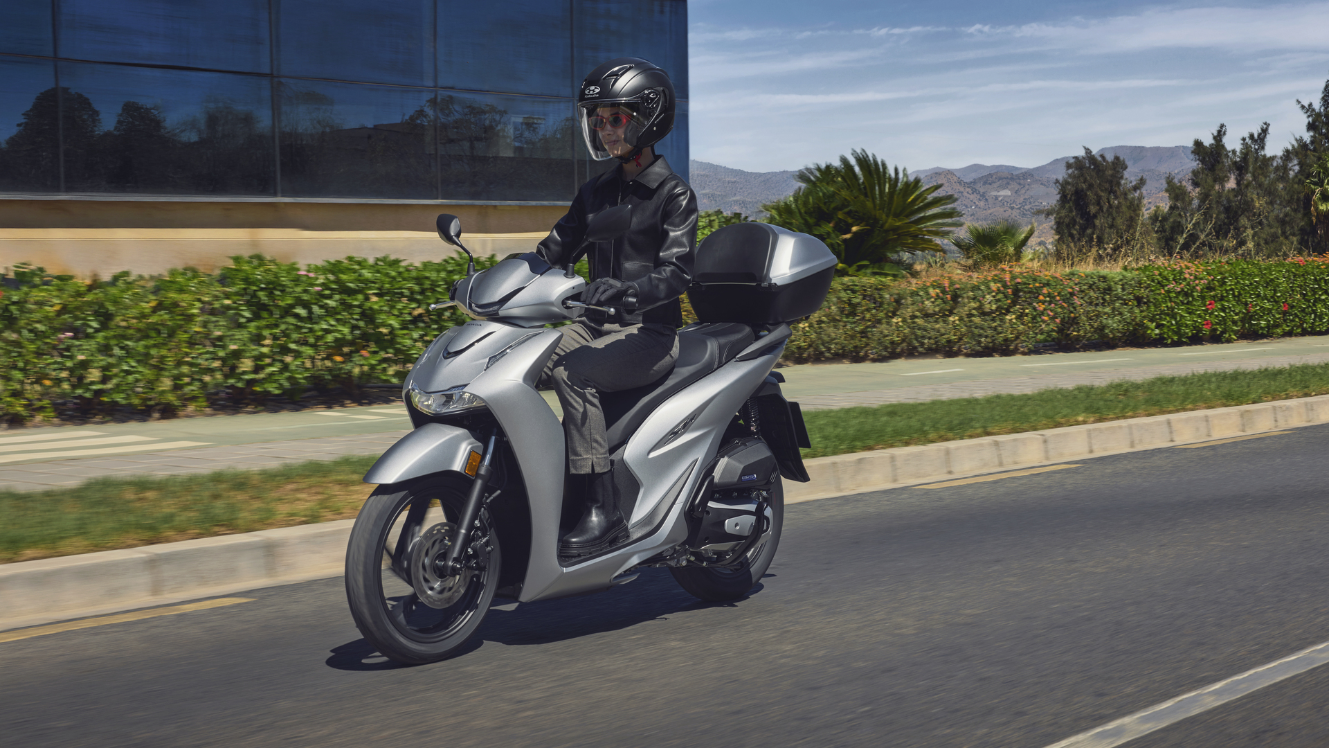A person riding a white Honda SH350i scooter on a sunny road, with green foliage and mountains visible in the background.