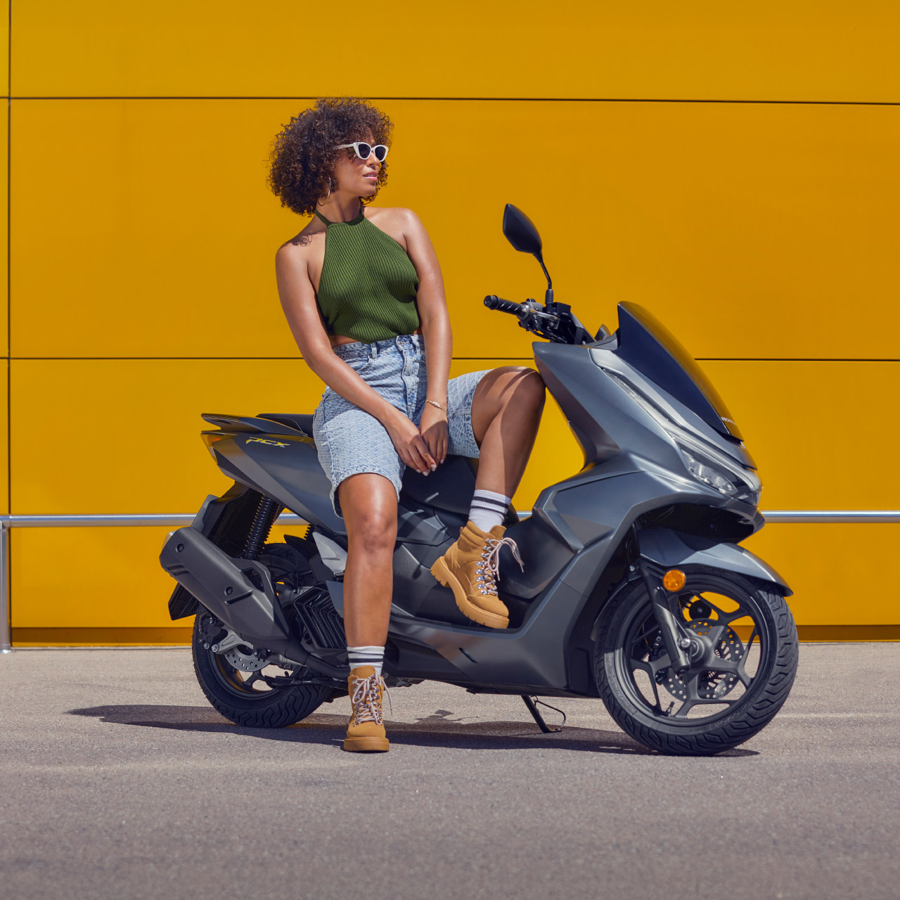 Eine Frau sitzt auf einem grauen Honda PCX 125 vor einer gelben Wand