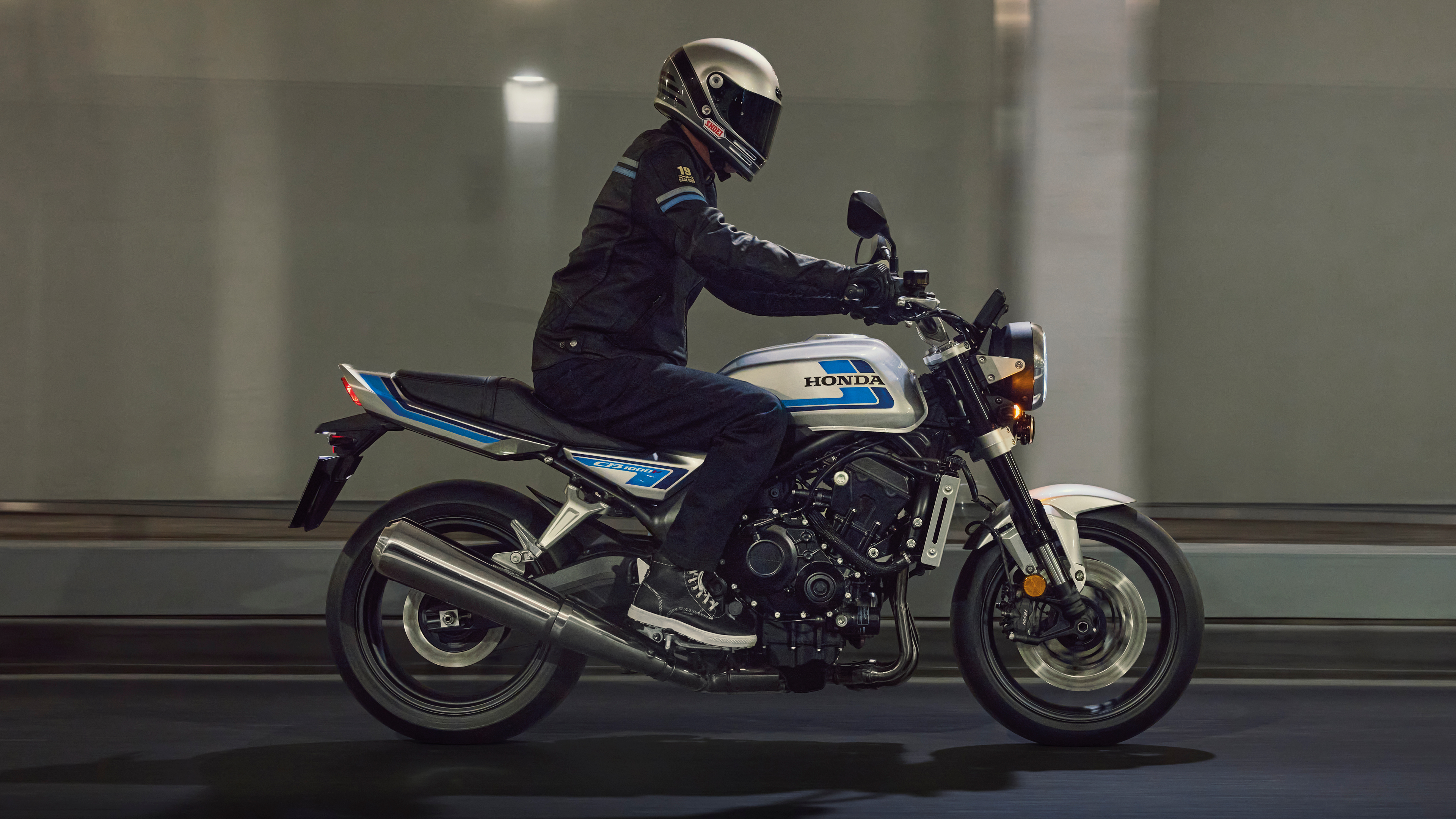 Ein Honda CB1000F Motorrad, silber und blau, in Bewegung in der Nacht. Der Fahrer trägt schwarze Schutzkleidung und einen silbernen Helm. Im Hintergrund sind reflektierende Schaufenster zu sehen, in denen sich das Licht der Straßenlaternen spiegelt. 